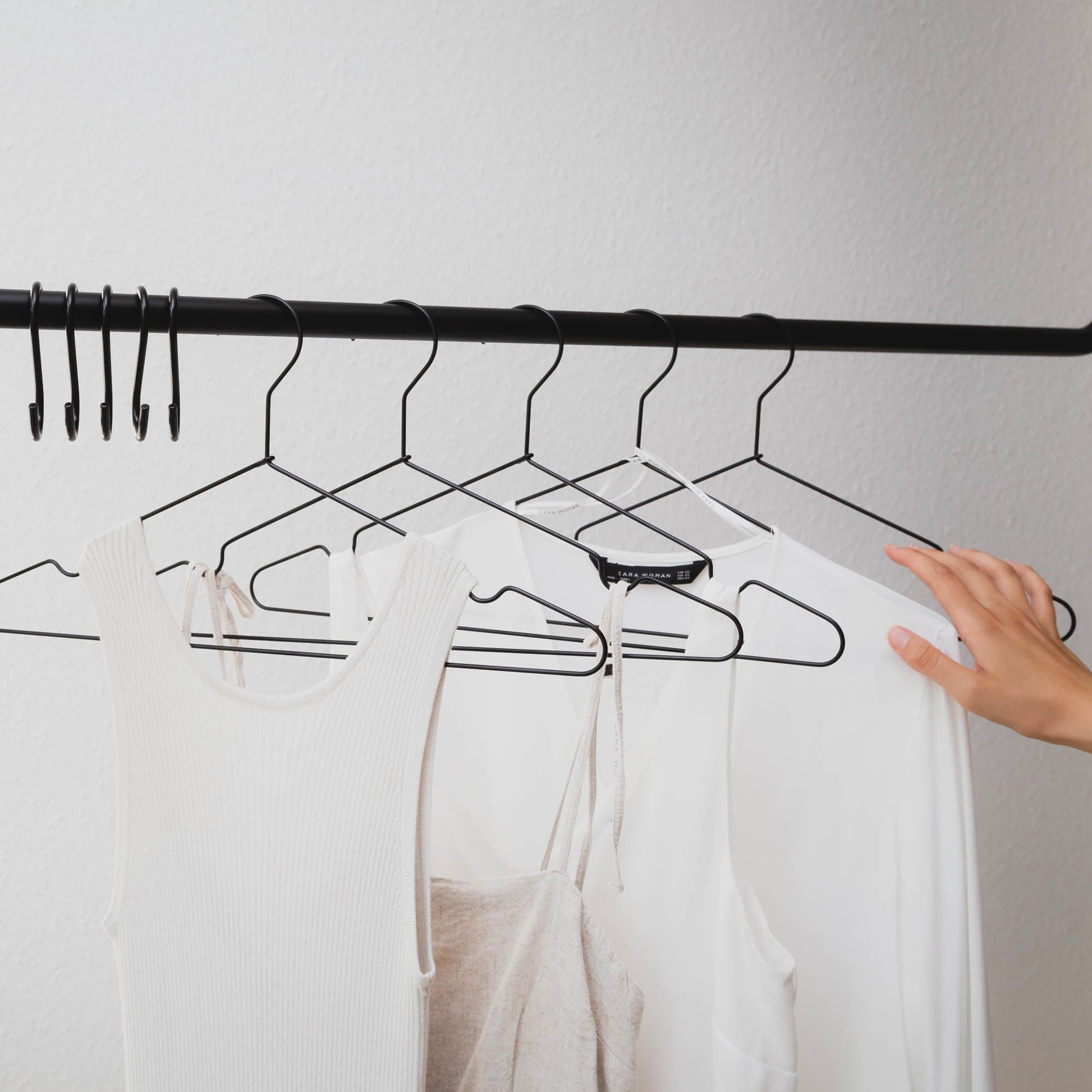 Eine Person ordnet mehrere Kleidungsstücke an einem Kleiderbügel aus stabilem Metall von Rod & Knot, der an einer schwarzen horizontalen Stange hängt. Die Kleidung umfasst ein weißes Tanktop, ein hellbeiges Kleid und ein weißes Langarmshirt. Der Hintergrund ist eine schlichte weiße Wand, die für platzsparende Aufbewahrung sorgt.